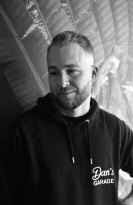 Porträtfoto von Daniel (Dan), dem Inhaber von Dan's Garage. Ein lächelnder Mann mit Vollbart und nach oben gestyltem Haar. Er ist in seiner Garage abgebildet, im Hintergrund ist die Werkstattumgebung leicht unscharf zu erkennen.