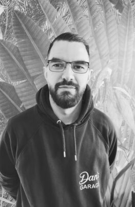 Porträt von Erwin , Mitarbeiter bei Dan's Garage. Er trägt eine Brille, hat dunkle Haare und einen gepflegten Vollbart. Er trägt einen dunklen Kapuzenpullover mit dem Logo von „Dan's Garage“ vor einem Hintergrund mit Palmenblatt-Motiv.