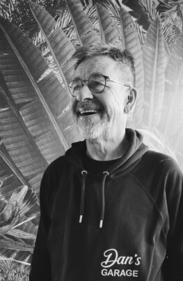 Porträtfoto von Pauli von Dan's Garage. Ein älterer Mann mit grauem Bart und Brille, der lachend zur Seite blickt. Er trägt einen schwarzen Kapuzenpullover (Hoodie) mit dem weißen Logo „Dan's GARAGE“. Der Hintergrund zeigt eine Schwarz-Weiß-Tapete mit großen Palmenblättern.